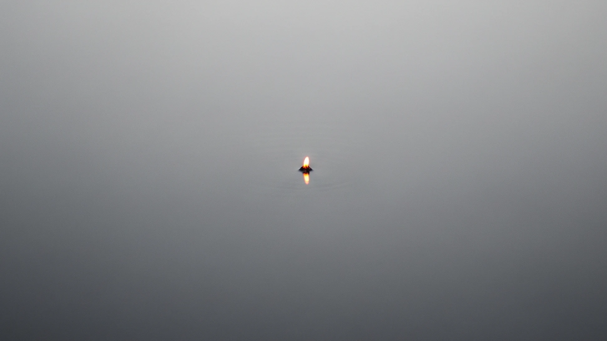 Minimalistische Szene einer kleinen, schwimmenden Kerze auf ruhiger, grauer Wasseroberfläche mit ihrer Spiegelung – Symbol für den Funken Hoffnung in der Stille der inneren Leere